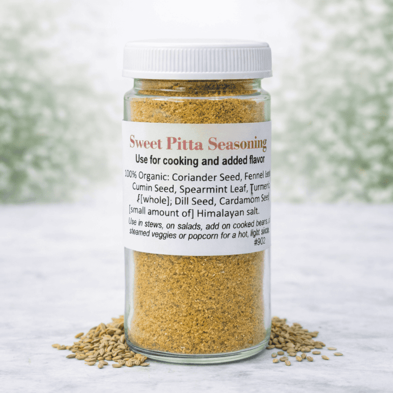A clear glass jar of Sweet Pitta Seasoning with a white lid and white label, placed on a light marble surface with loose seasoning scattered around the base. The background is a soft, blurred white and green outdoor-style backdrop.